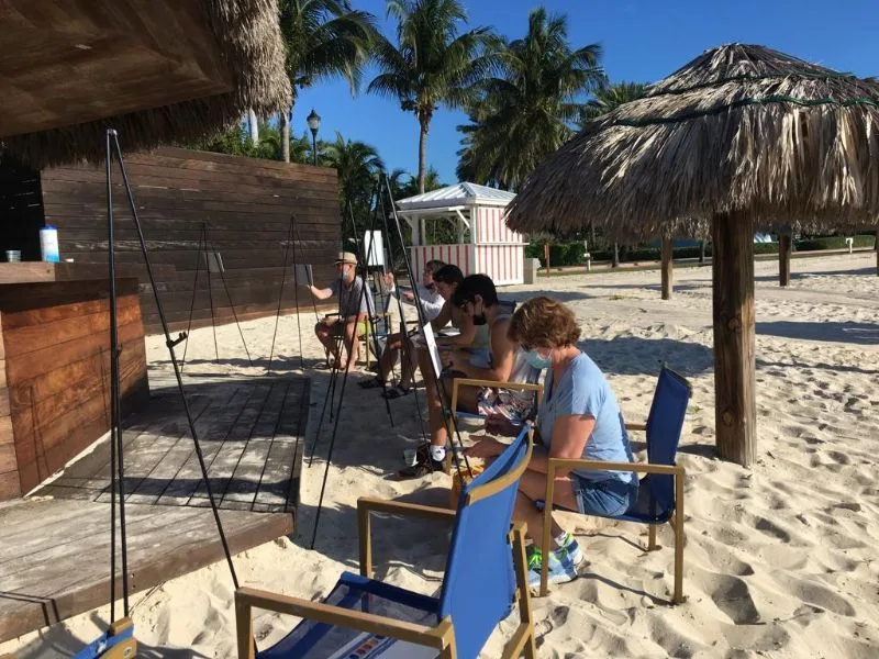 Guests painting at easels under thatched umbrellas on a Bimini beach